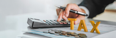 Een hand die een pen beet houdt en tegelijkertijd iets intoetst op de rekenmachine met het woord TAX ervoor en wat geld