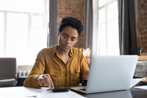 Dame met kort zwart krullend haar achter laptop en iets aan het uitrekenen op rekenmachine